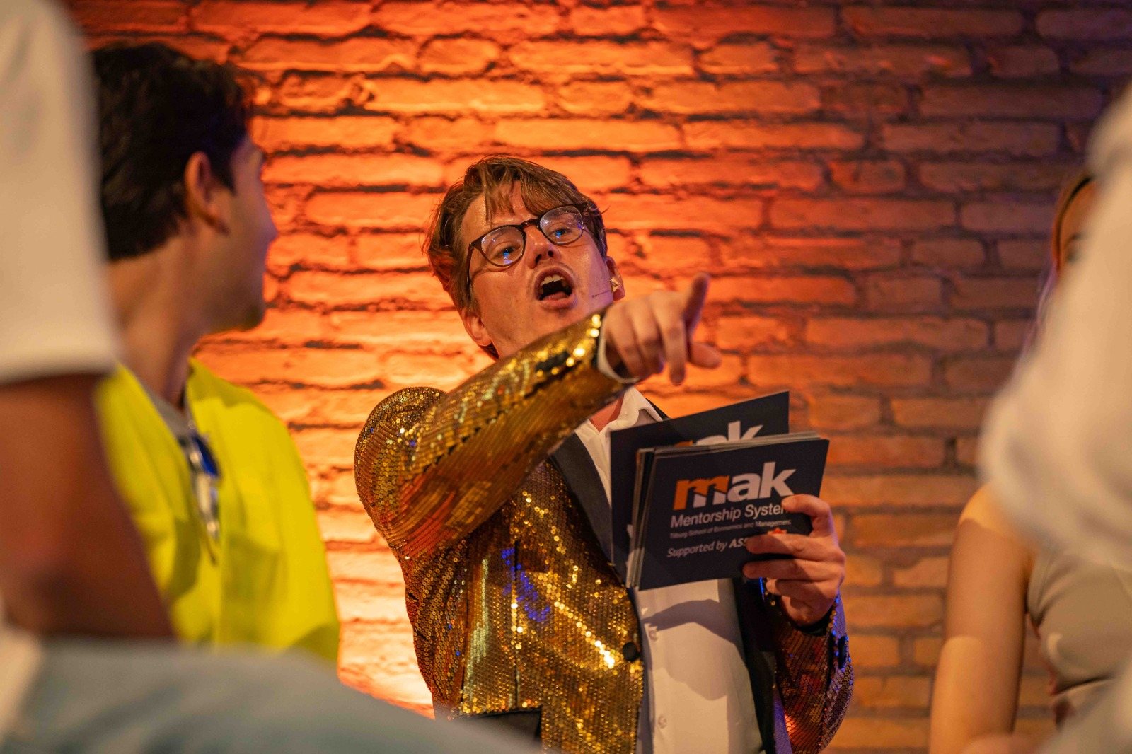 Jerre Maas als quizmaster in een glitterjasje wijst een deelnemer aan tijdens een quiz van MAK
